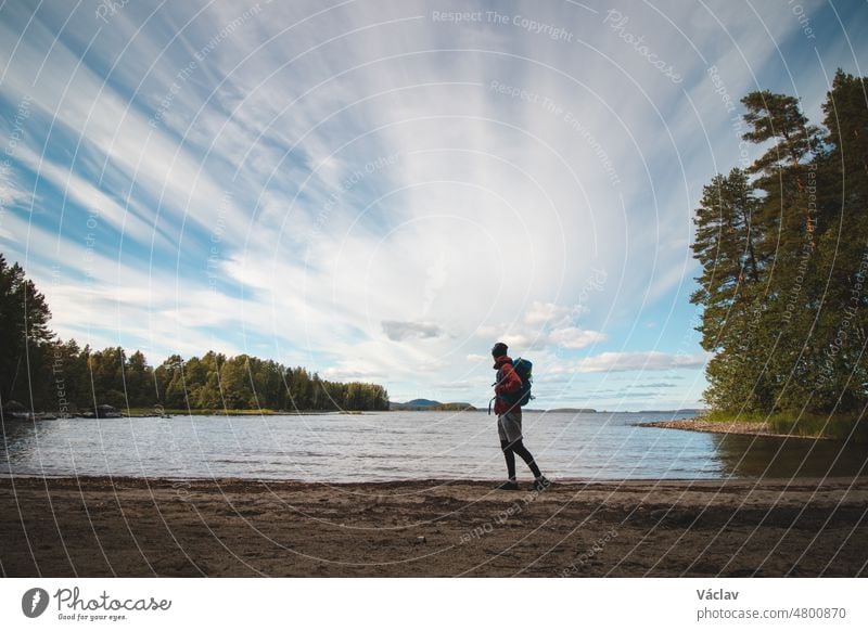 Wanderer mit Jacke und Rucksack geht am Strand entlang und beobachtet den See Jatkonjarvi bei Sonnenuntergang im Koli-Nationalpark in Ostfinnland. Mann im Alter von 24 Jahren in Sportkleidung. Aktiver Lebensstil