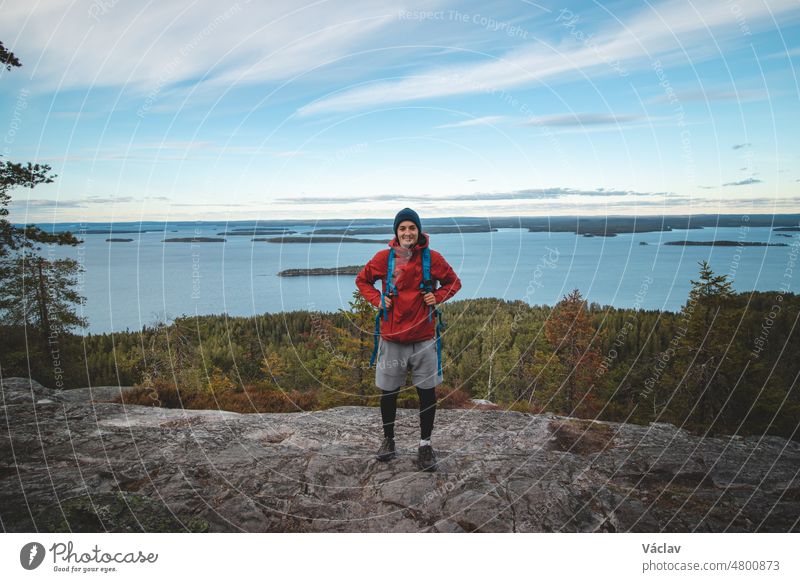 Lachender Rucksacktourist auf einem Felsen mit dem See Jatkonjarvi im Koli-Nationalpark, Ostfinnland. Der Mann, 24 Jahre alt, lächelt natürlich. Aktiver Lebensstil