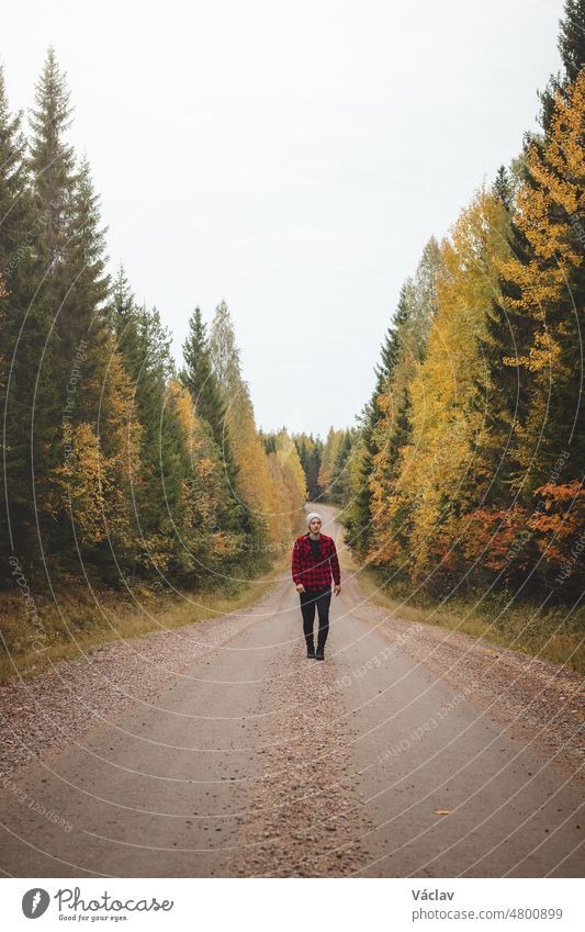 Ein 23-jähriger Mann in einem rot-schwarz karierten Hemd geht einen schaumigen Weg entlang, der von schönen, herbstlich gefärbten Laubbäumen umgeben ist. Genießen Sie den Moment. Region Kainuu, Finnland
