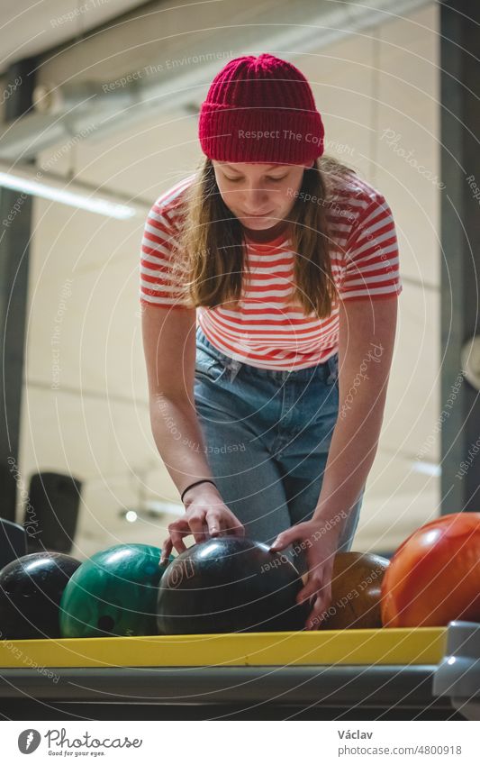 Eine junge Amateur-Bowlerin mit roter Mütze und gepunktetem T-Shirt nimmt die ideale Kugel mit dem richtigen Gewicht und bereitet sich innerlich auf ihren Wurf vor. Konzentration