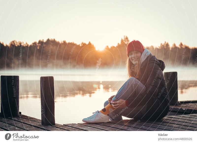 Junge Brünette mit einem schönen, realistischen Lächeln sitzt auf einer Holzbrücke an einem See bei Sonnenaufgang. Roter Hut mit schwarzem Mantel und weißen Schuhen. Modischer Stil. Paltamo, Finnland. Entdeckung von Skandinavien