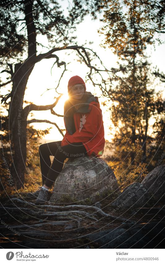 schöne brünette Reisende sitzt auf dem Gipfel eines Hügels auf einem Felsen und ruht sich nach einem anstrengenden Aufstieg aus. Sonnenuntergang auf dem Gipfel eines Berges in Vuokatti, Region Kainuu, Finnland. Natürliches Lächeln eines Mädchens in ihren 20ern