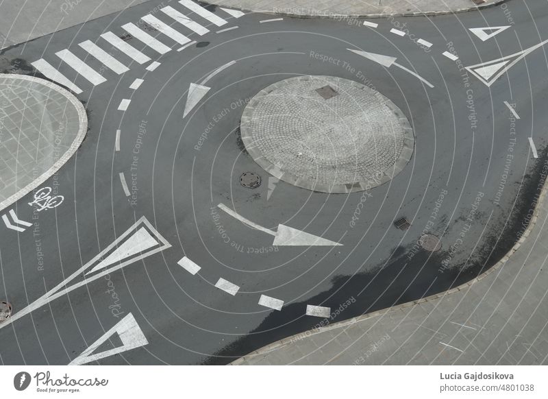 Blick von oben auf einen leeren Kreisverkehr mit weißen Pfeilen, Zebrastreifen und anderen Straßensignalen oder -markierungen in einer europäischen Stadt. Der graue Asphalt ist nach dem Regen teilweise nass.