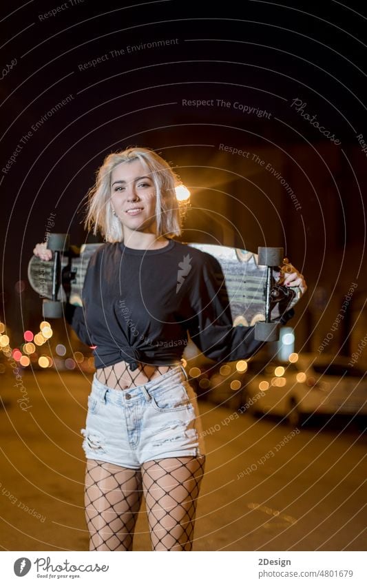 junge hübsche Frau in Shorts, die auf der Straße steht und ein Skateboard hält, nachts in der Stadt Vorderansicht sinnlich schauende Kamera Beteiligung
