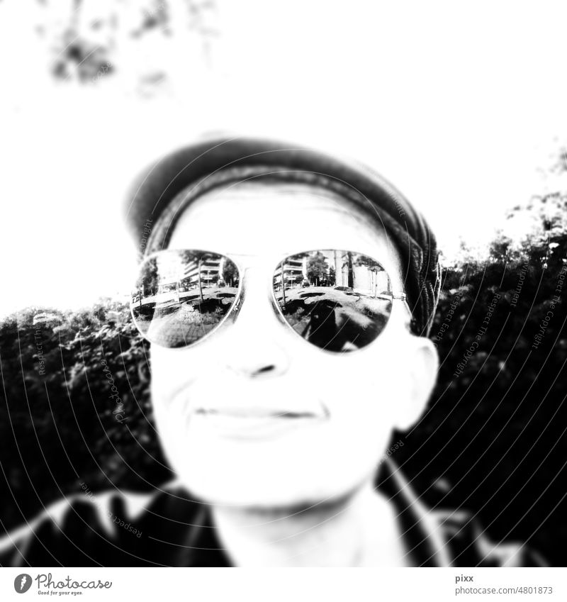 Freundlich lächelnde Person mit Sonnenbrille in der sich urbanes Leben spiegelt. Trägt Schiebermütze. Schwarz-Weiß Foto. Quadratisch Schwarzweißfoto Brille