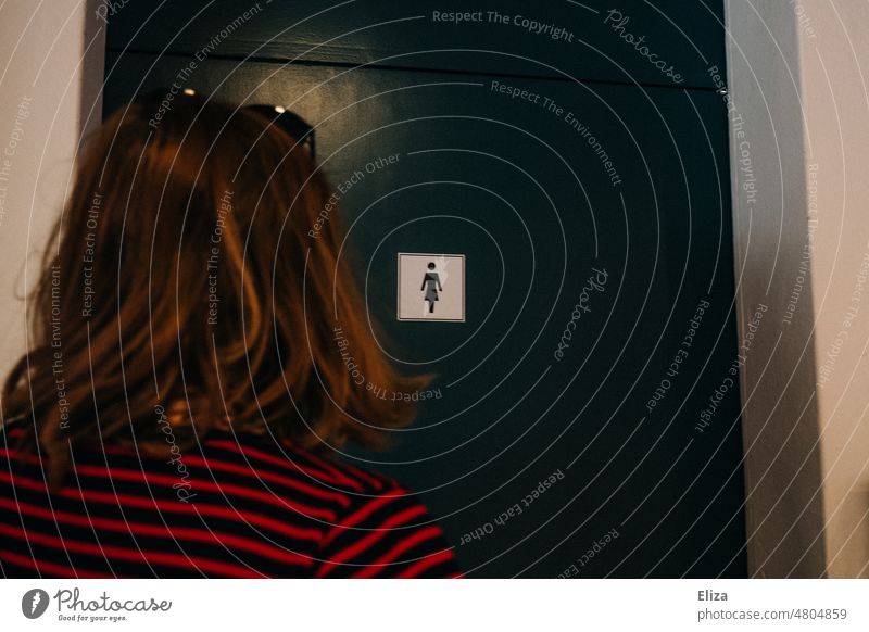 [hansa BER 2022] Eine Frau steht vor einer Türe mit dem Symbol für Damentoilette Toilette Piktogramm WC Frauentoilette rot-schwarz gestreift blau Gender