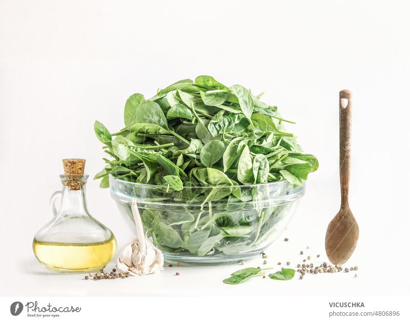 Kochgeschirr mit grünem Blattspinat in Glasschale, Olivenöl und Holzlöffel auf weißem Hintergrund Essen zubereiten Einstellung Spinat Schalen & Schüsseln