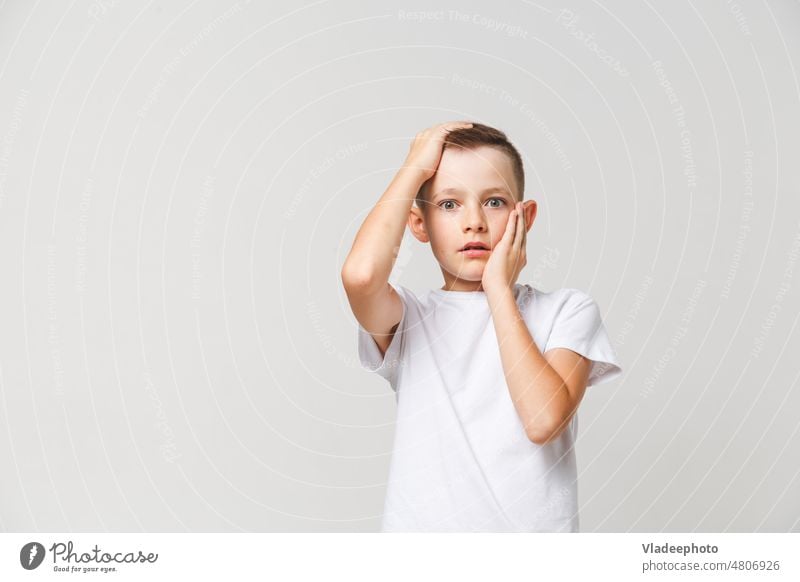 Verängstigter Junge in weißem T-Shirt mit beiden Händen auf dem Kopf auf grauem Hintergrund entsetzt verängstigt furchtbar Kind ängstlich Angst Person jung
