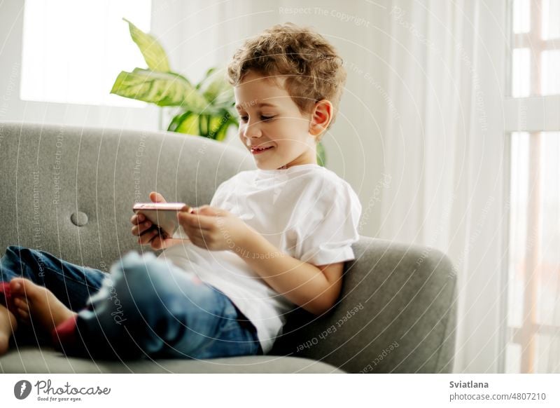 Ein fröhlicher Junge mit einem Telefon in der Hand sitzt auf einem Stuhl und spielt ein Spiel oder schaut Cartoons. Smartphone Spielen wenig Sitzen heimwärts