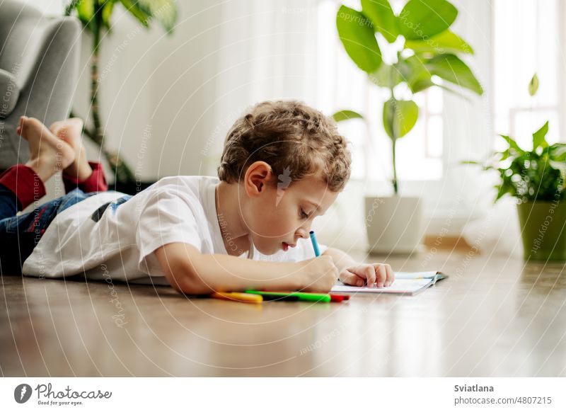 Ein kleiner Junge zeichnet, liegt auf dem Boden und hat Spaß. Glückliche Kindheit, positive Emotionen. Seitenansicht, Platz für Text Zeichnung heimwärts Stock