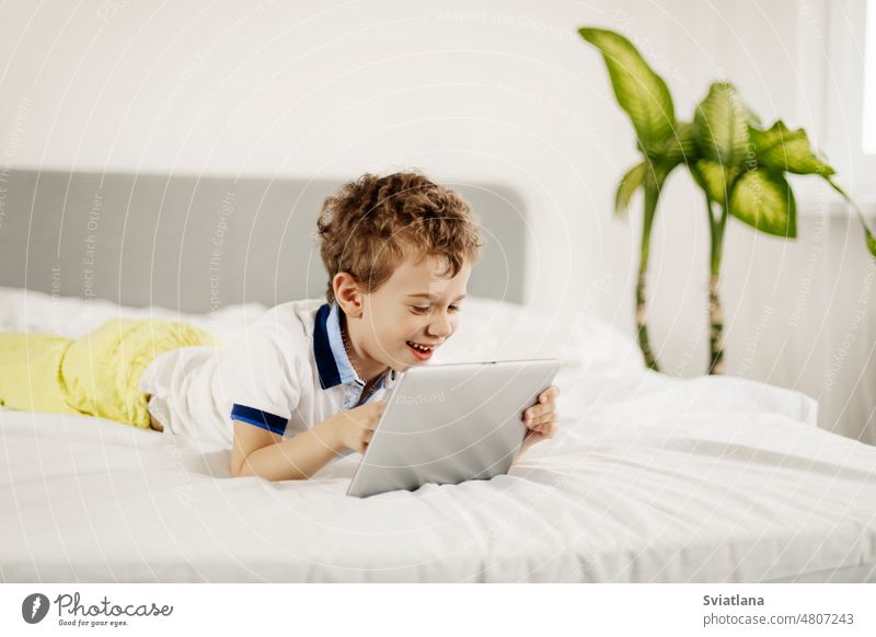 Lockenköpfiger Junge liegt auf dem Bett im Schlafzimmer und spielt Computerspiele auf einem Tablet Tablette Lügen Technik & Technologie Glück Kind