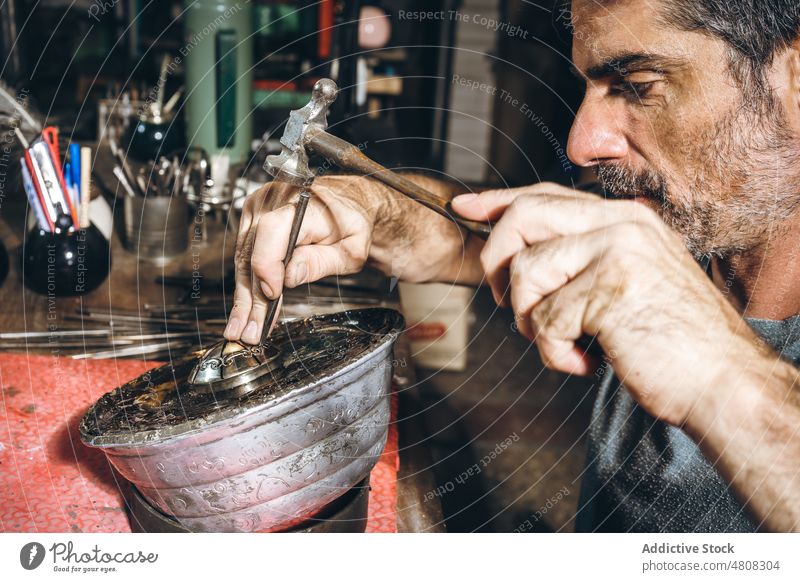 Männlicher Silberschmied, der ein Ornament auf ein silbernes Accessoire stanzt Mann Bowle Hammer Werkstatt professionell Kunstgewerbler männlich reif