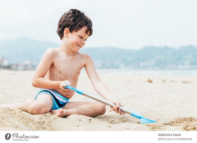 Fröhlicher kleiner Junge gräbt Sand am Strand Graben Sommer Urlaub Lächeln schaufeln Wochenende froh Kind Küste sich[Akk] entspannen Ufer knien Kindheit ruhen