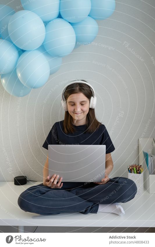 Lächelndes Teenager-Mädchen mit Kopfhörern, das auf einem Tisch sitzt und einen Laptop benutzt benutzend Internet zuschauen Video positiv Beine gekreuzt