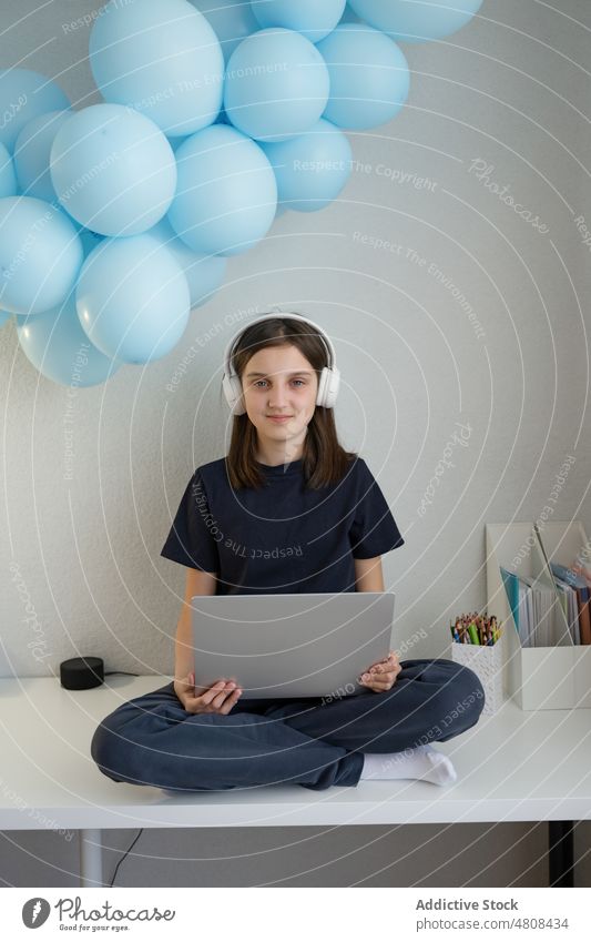 Lächelndes Teenager-Mädchen mit Kopfhörern, das auf einem Tisch sitzt und einen Laptop benutzt benutzend Internet zuschauen Video positiv Beine gekreuzt