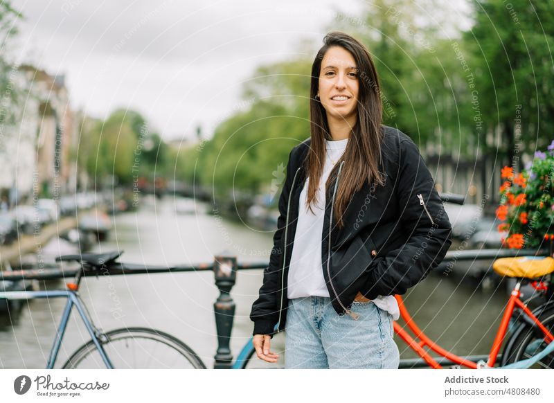 Glückliche ethnische Frau lächelt, während sie auf einer Fußgängerbrücke an einem bewölkten Tag steht Lächeln Brücke Großstadt Kanal Fahrrad Reisender Stil