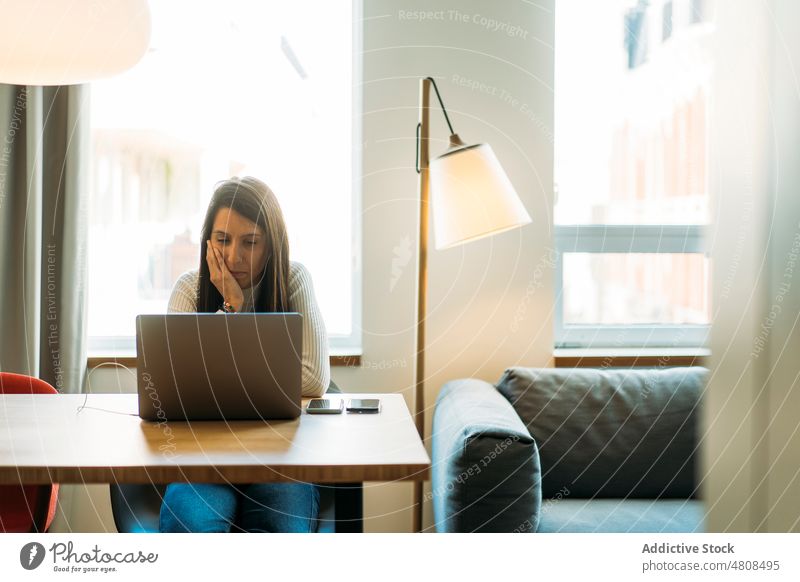 Fokussierte junge ethnische Frau am Tisch sitzend mit Laptop und Smartphones freiberuflich nachdenklich online Hand auf die Wange Arbeit Anschluss abgelegen