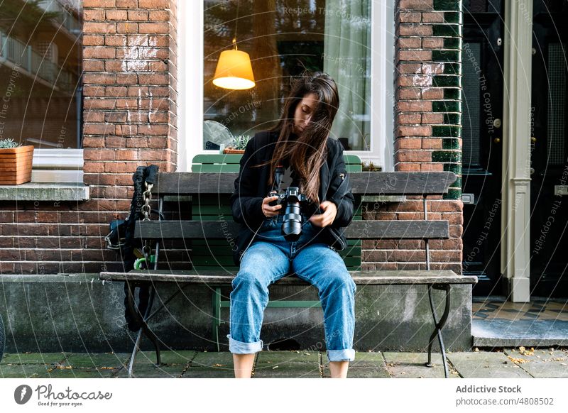 Seriöse junge ethnische Dame mit Fotokamera auf einer Bank in einer Stadtstraße sitzend Frau Fotoapparat Fotograf Fotografie Straße prüfen Reisender Gebäude