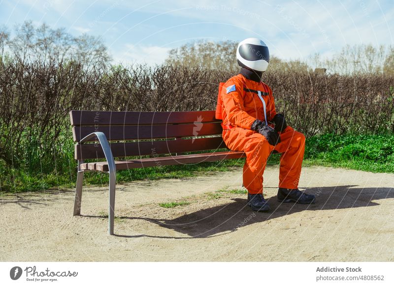 Unbekannter Raumfahrer mit Schutzhelm und Kostüm sitzt auf einer Bank Astronaut Park Kosmonaut Natur futuristisch behüten Raumanzug warten Sicherheit Tracht