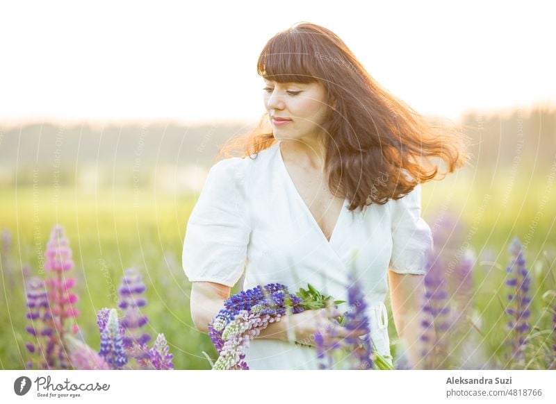 Schöne Frau im weißen Sommerkleid genießt die sommerliche Natur. Pflücken bunte Blumen, atmen frische Luft und Blumenduft, zu Fuß in sonnigen Feld von Lupinen, Wind in den Haaren. Glück Konzept