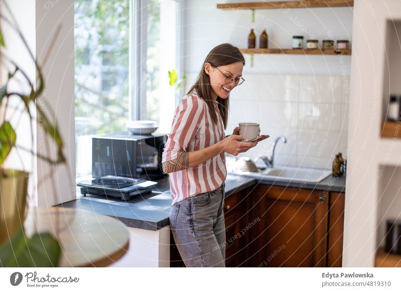 Junge Frau benutzt ihr Smartphone, während sie in ihrer Küche zu Hause steht Handy Technik & Technologie online Internet per Telefon Mitteilung häusliches Leben