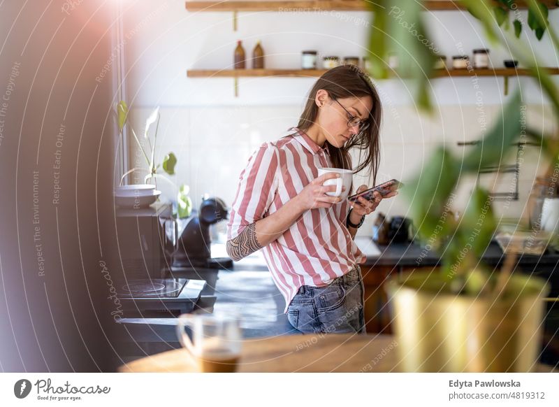 Junge Frau benutzt ihr Smartphone, während sie in ihrer Küche zu Hause steht Handy Technik & Technologie online Internet per Telefon Mitteilung häusliches Leben