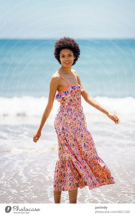 Glückliche schwarze Frau im Sommerkleid, die auf dem Sand eines tropischen Strandes spazieren geht. Afro-Look Frisur Spaziergang sich[Akk] entspannen Küste