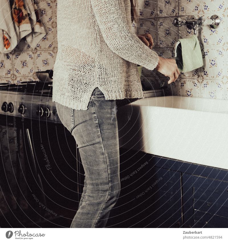 Frau in Jeans und weißem Strickpullover am Waschbecken einer einfachen Küche mit Gasherd und bunten Kacheln Herd Wasserhahn Abwasch Küchenarbeit Hausarbeit