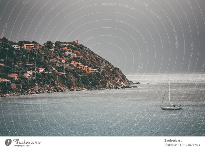 Segelyacht liegt vor düsterem Himmel in einer Bucht auf Sardinien vor Anker, im Hintergrund Hügel und Ferienhäuser Segelboot Boot Ankern Wolken