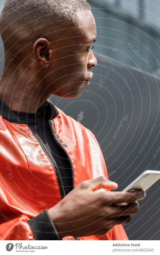 Fokussierter schwarzer Mann, der sein Smartphone benutzt, während er sich auf der Straße an eine Wand lehnt Nachricht Großstadt trendy benutzend Stil ernst