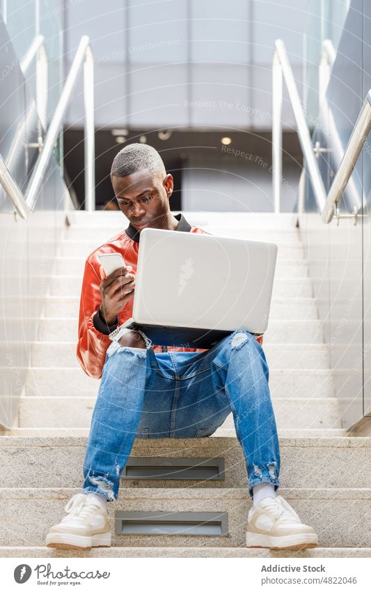 Selbstständiger schwarzer Mann mit Smartphone und Laptop auf der Treppe freiberuflich abgelegen Straße Großstadt multitask männlich jung Afroamerikaner ethnisch
