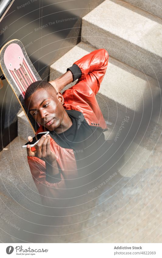 Trendiger schwarzer Mann, der eine Sprachnachricht auf seinem Smartphone sendet und sich auf ein Skateboard stützt Sprachmeldung sich[Akk] entspannen Audio