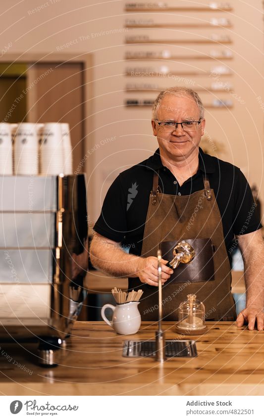 Senior Barista mit Filter in der Hand lächelnd im Kaffeehaus Mann Kaffeemaschine Portafilter Lächeln positiv Abfertigungsschalter Café Porträt Arbeit