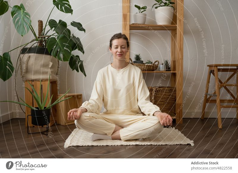 Friedliche Frau meditiert in Lotus-Yoga-Pose zu Hause Lotus-Pose padmasana meditieren Augen geschlossen Zen Stressabbau üben Asana Wellness Windstille jung