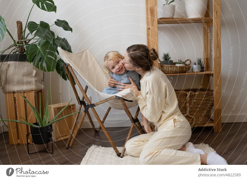 Zufriedene Mutter, die auf dem Boden sitzt und ihr Kind zu Hause umarmt Frau Kuss Stuhl Liebe Glück Mutterschaft Umarmen positiv Appartement bezaubernd