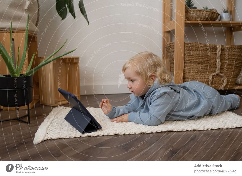 Niedliches Kleinkind liegt auf dem Boden und schaut sich ein Video auf dem Tablet an Kind zuschauen Tablette Lügen Stock neugierig Interesse achtsam Karikatur