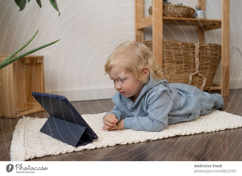 Niedliches Kleinkind liegt auf dem Boden und schaut sich ein Video auf dem Tablet an Kind zuschauen Tablette Lügen Stock neugierig Interesse achtsam Karikatur