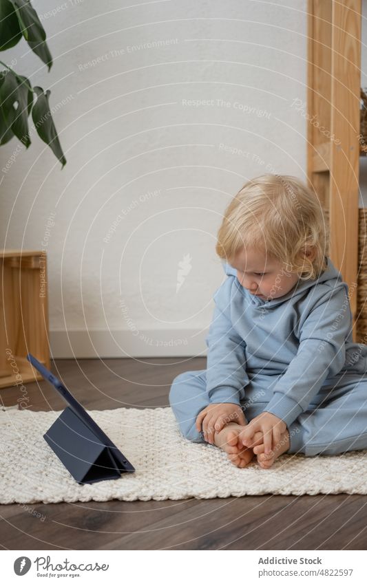Niedliches Kleinkind sitzt auf dem Boden und schaut sich ein Video auf dem Tablet an Kind zuschauen Tablette Stock neugierig Interesse achtsam Karikatur