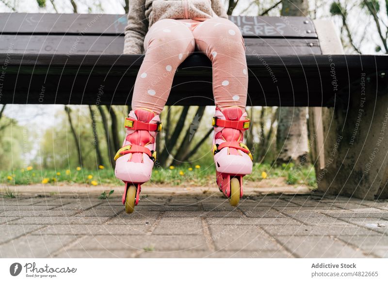 Anonymes süßes Kind in Rollschuhen, das sich auf einer Bank im Park ausruht Rollerskate ruhen Mädchen aktiv Hobby bezaubernd Aktivität Schlittschuh lässig