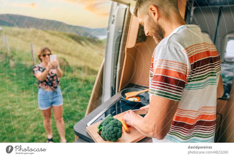 Junger Mann kocht Gemüse in einem Wohnmobil, während seine Frau ein Foto von ihm mit ihrem Handy macht jung Essen zubereiten Ehefrau Bild Mobile Veganer Freund