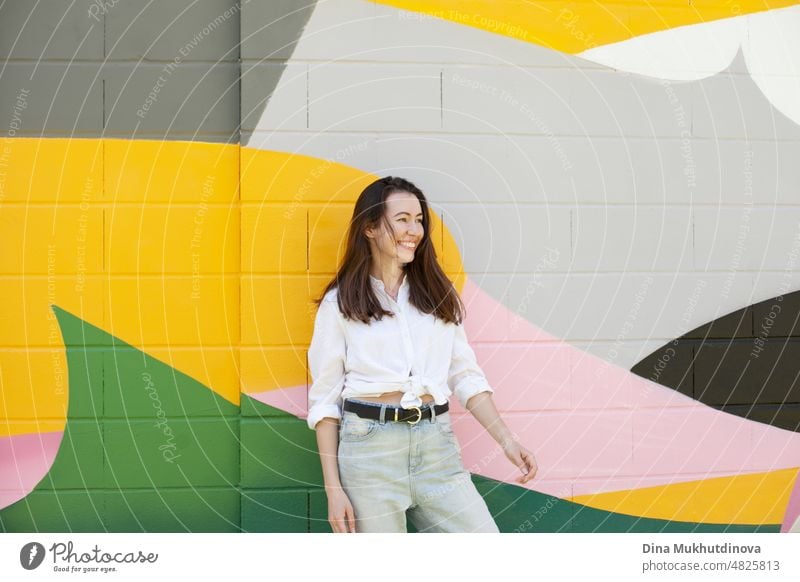 Junge Frau in weißem Hemd und hellblauen Jeans stehen in der Nähe von bunten lebendigen Wand. Candid Lifestyle Sommer Stil Porträt. schön Großstadt Glück