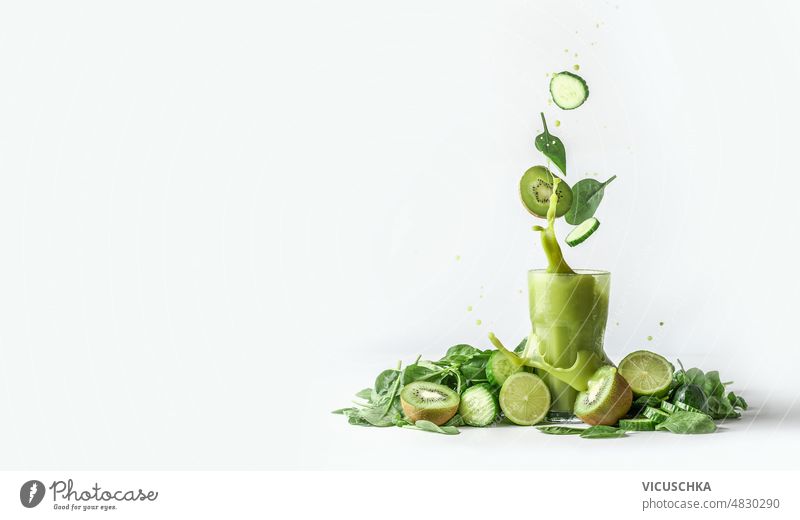 Grüner Smoothie spritzt im Glas und fliegende Zutaten auf weißem Hintergrund mit Haufen von grünem Obst und Gemüse Geplätscher Salatgurke Kiwi Spinatblätter