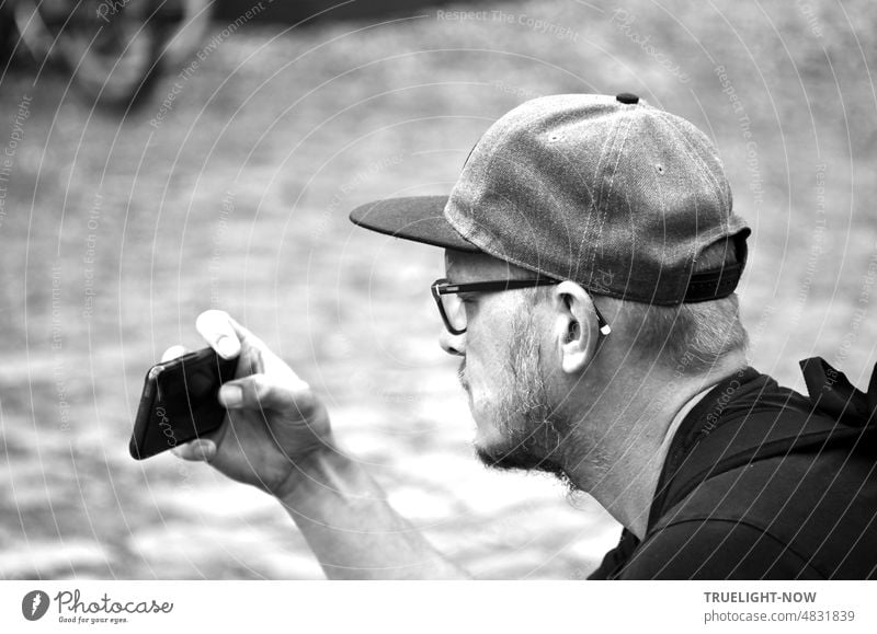 [Hansa BER 2022] Portrait Junger Mann mit Brille und Basecap blickt konzentriert auf sein Smartphone, das er zum Fotografieren in der rechten Hand vor sich hält. b/w schwarz-weiss