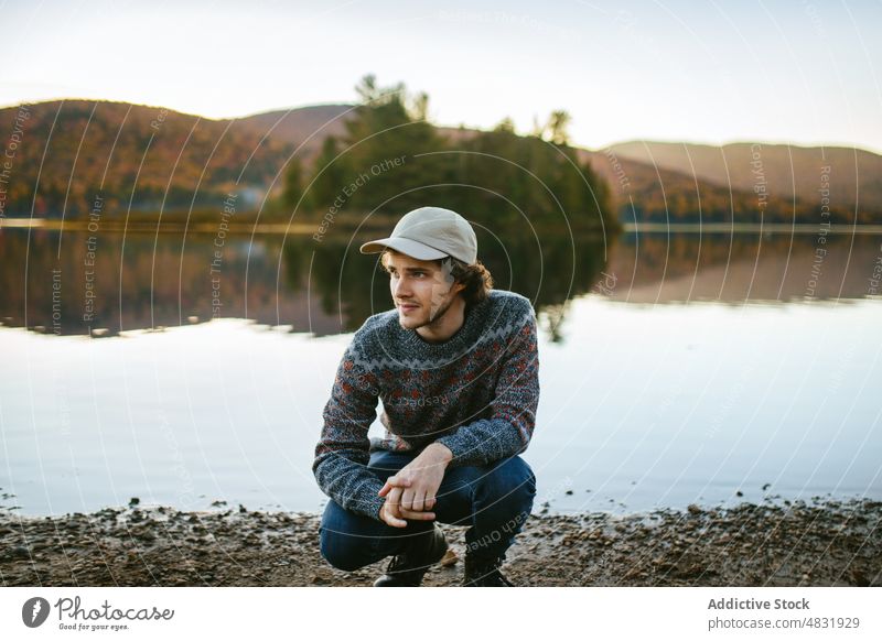 Stilvoller Reisender, der an einem Herbsttag am Seeufer sitzt Mann Natur bewundern Ausflug Ufer positiv fallen Hügel reisen malerisch männlich jung Pullover