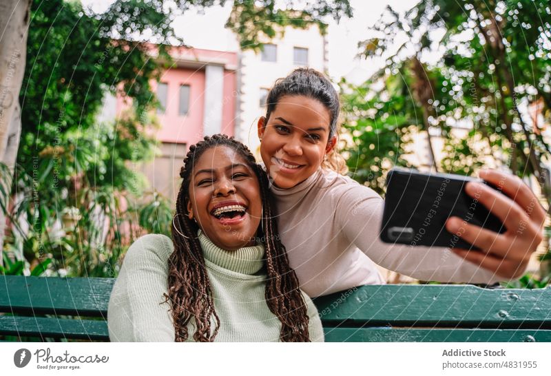 Glückliche diverse Freundinnen nehmen Selfie auf Bank Frauen Park Smartphone Lachen Zusammensein Lächeln vielfältig rassenübergreifend multiethnisch schwarz