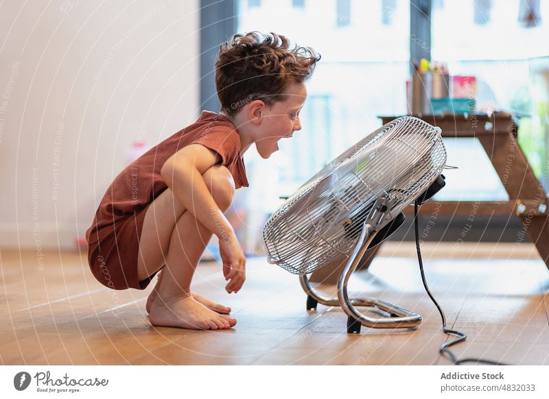 Kleiner Junge sitzt zu Hause auf dem Boden und spielt mit einem Ventilator Schlag spielen Spiel jung genießend blasend Spielen Ausdruck Spaß Offener Mund