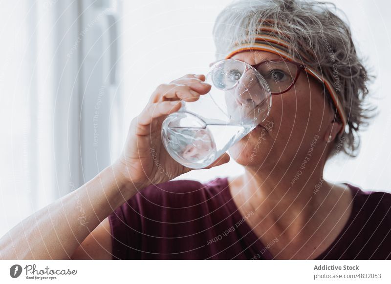 Ältere Frau trinkt Wasser beim Fitnesstraining trinken Training Pause heimwärts Morgen Hydrat Durst älter Haarband Senior gealtert ruhen Fenster Wohlbefinden