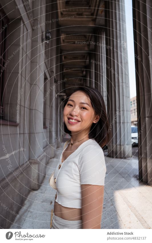 Zufriedene junge Asiatin steht in der Nähe eines Gebäudes mit Säulen im Sonnenlicht Frau Lächeln Durchgang Urlaub Sightseeing Glück Tourist Spalte positiv Stil