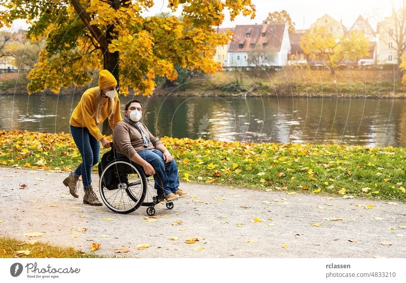 Frau und ihr Freund mit Gesichtsmaske im Rollstuhl machen einen Spaziergang durch den Park an einem Fluss und genießen den Herbst während der Covid-19-Pandemie