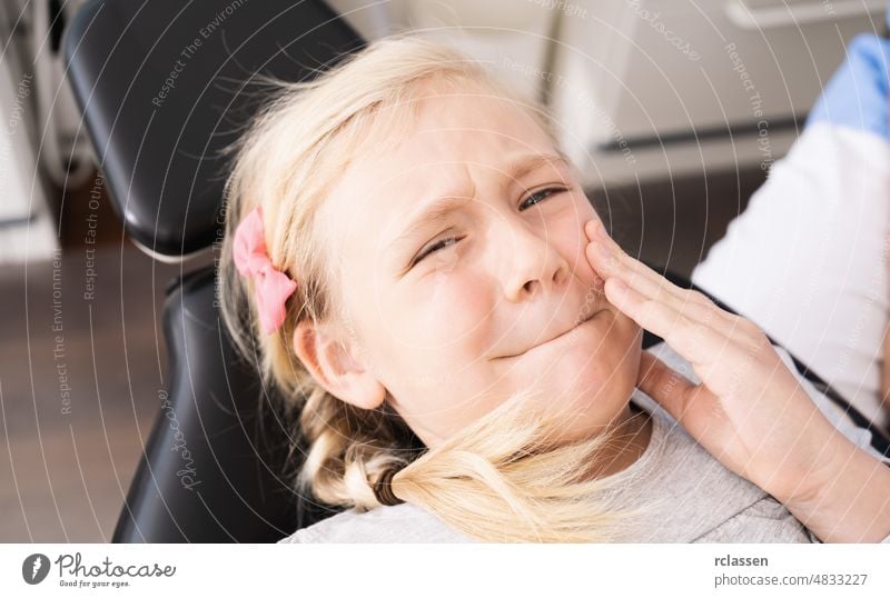 Kleines Kind beim Zahnarzt in bequemen Stuhl berührt den Mund mit der Hand mit schmerzhaften Ausdruck wegen Zahnschmerzen oder Zahnerkrankung auf die Zähne. Zahnarzt Konzept Bild